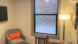 Ein Zimmer mit zwei grauen Sesseln, davon einer mit einem orangefarbenen Kissen, einem kleinen schwarzen Hocker, einer Stehlampe und einem Fenster mit Blick auf eine Ziegelwand.