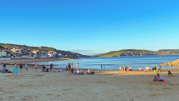 Een strand met mensen die zonnebaden en wandelen, een heldere lucht, en gebouwen op de achtergrond.