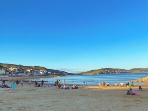 Une plage où l’on voit des gens se prélasser au soleil et se promener, un ciel dégagé et des bâtiments en arrière-plan.