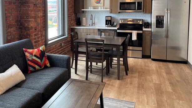 A modern kitchen with wooden cabinets, stainless steel appliances, a dining area with a table and chairs, and a gray sofa with a colorful pillow.