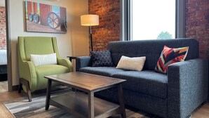 A modern living room with a green armchair, a dark grey sofa, and a wooden coffee table. A painting of wheels hangs on the brick wall, and a floor lamp is positioned near the sofa.