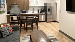 A compact kitchen with wooden cabinets, a microwave, a stove, and a refrigerator. There is a dining table with chairs and a sofa with a patterned pillow.