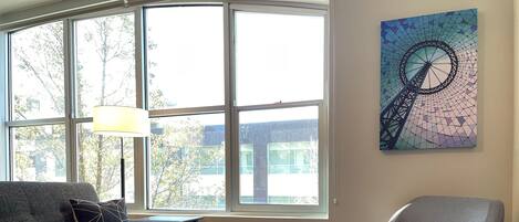 A living room with a sofa, armchair, and coffee table, large windows with a view of trees and buildings, and a wall-mounted abstract art piece.