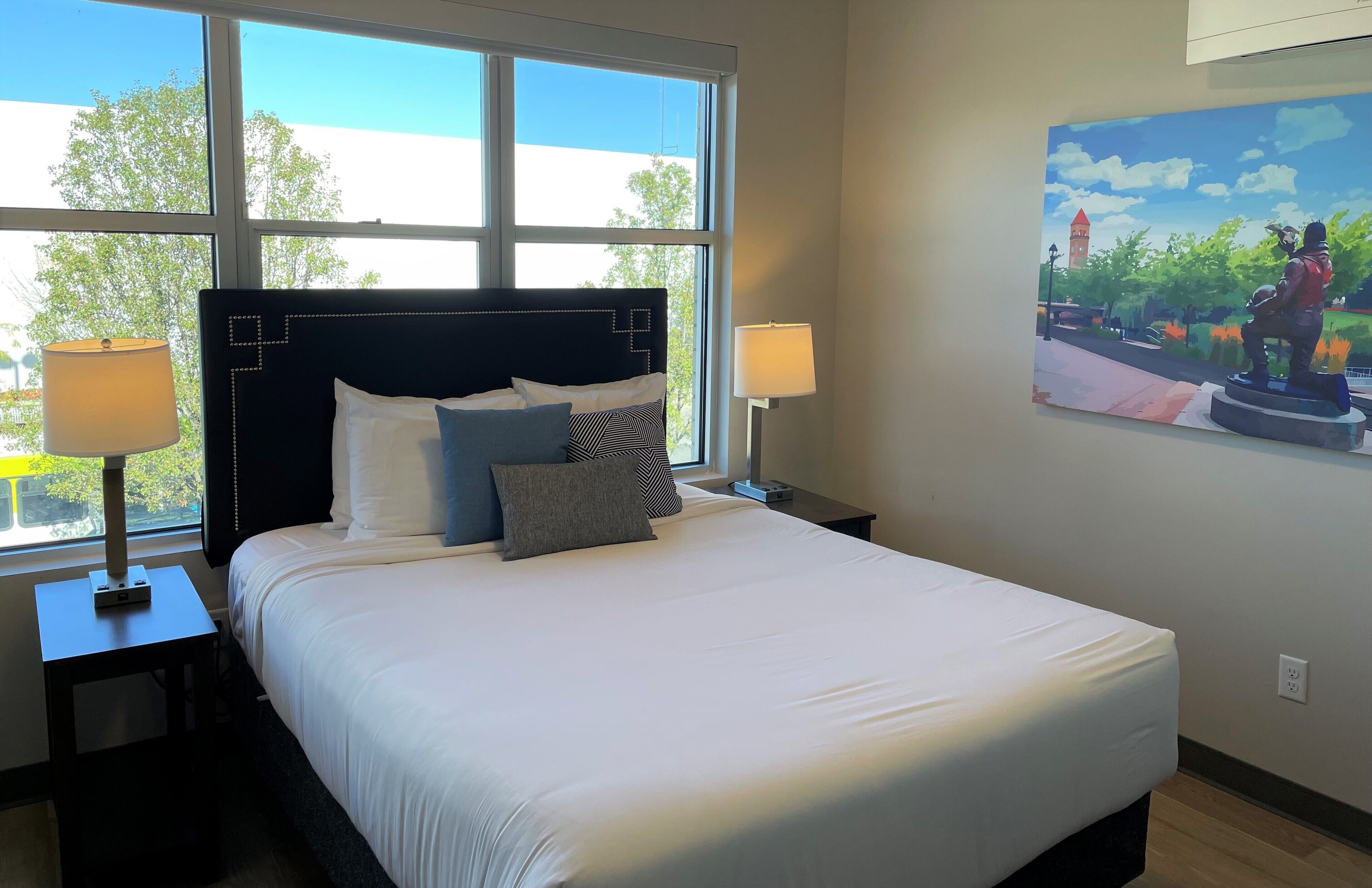 A hotel room with a large bed, two bedside tables with lamps, a painting on the wall, and a window with a view of trees and a building.