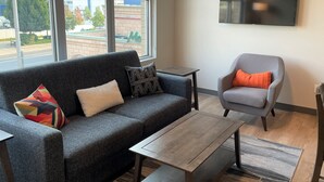 Ein modernes Wohnzimmer mit einem grauen Sofa, einem grauen Sessel und einem Couchtisch. Ein Fernseher ist an der Wand befestigt und große Fenster bieten Blick auf die Stadt.
