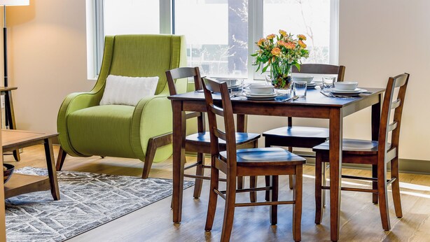 Un coin repas avec une table en bois dressée pour quatre personnes, entourée de chaises en bois. Un fauteuil vert est placé à gauche de la table, et un vase rempli de fleurs se trouve sur la table.