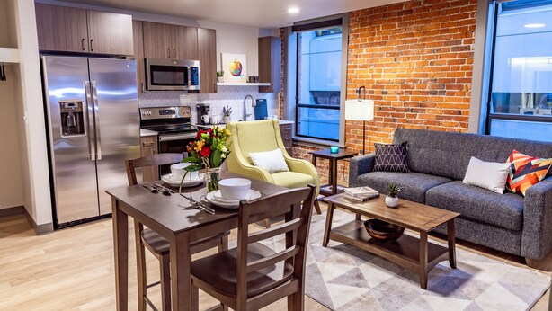 A modern kitchen with stainless steel appliances, a dining area with a wooden table and chairs, a yellow armchair, and a grey sofa with colorful cushions.