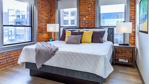 A modern bedroom with a brick wall, large windows, a bed with a white quilt, and two bedside lamps.