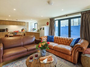 A modern living room with a leather sofa, a coffee table with food and drinks, and a kitchen in the background. - Coach House - UK32202 (Burton Bradstock, near Bridport)