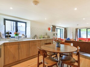 A modern kitchen with wooden cabinets, a dining table with chairs, a refrigerator, and a window with curtains.