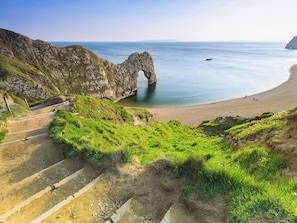 Paisagem costeira com um arco natural de rocha, uma praia arenosa e uma trilha de terra que leva ao arco.
