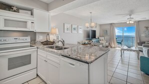 A modern kitchen with a built-in microwave, oven, and dishwasher. There is a large island with a sink and a chandelier above. The room has a large window with a view of the ocean. - Fourwinds 803-Cozy Beachfront condo-Hall Bunks-less crowded beaches-Epic Views (Orange Beach)