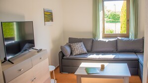 A modern living room with a flat-screen TV, a sofa with cushions, a small table with a cup and a notebook, and a window with curtains.