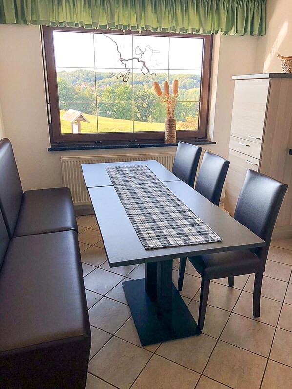 Un comedor con una mesa y sillas, vistas del exterior a través de una ventana, y un radiador debajo de la ventana.