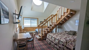 A cozy room with a wooden staircase, a bed with a patterned quilt, a dining table with chairs, and a window with blinds. - LAKEFRONT FISHING LODGE RETREAT SLEEPS 50! BOAT RENTALS! WORLD CLASS FISHING! (Panguitch)