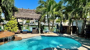 A swimming pool with a thatched roof structure, surrounded by palm trees and lounge chairs.