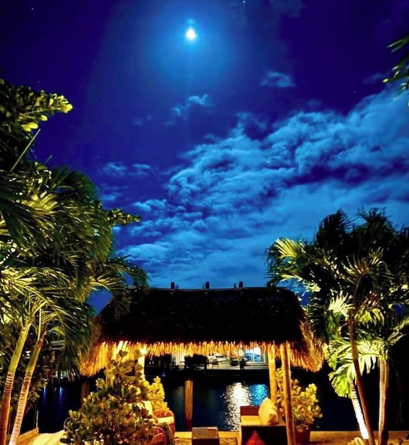 Una vista nocturna de un resort con una estructura de techo de paja, palmeras y una masa de agua.