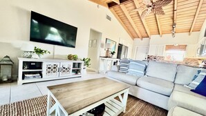 A living room with a flat-screen TV, a wooden coffee table, a grey sofa, and a kitchen in the background.