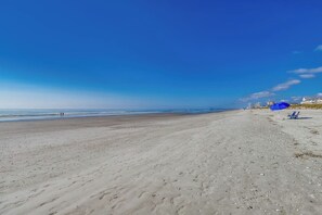 A wide sandy beach with footprints leading to the water, a blue umbrella, and a few beach chairs.