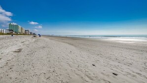 Una amplia playa de arena con huellas, un muelle a lo lejos y edificios a la izquierda.