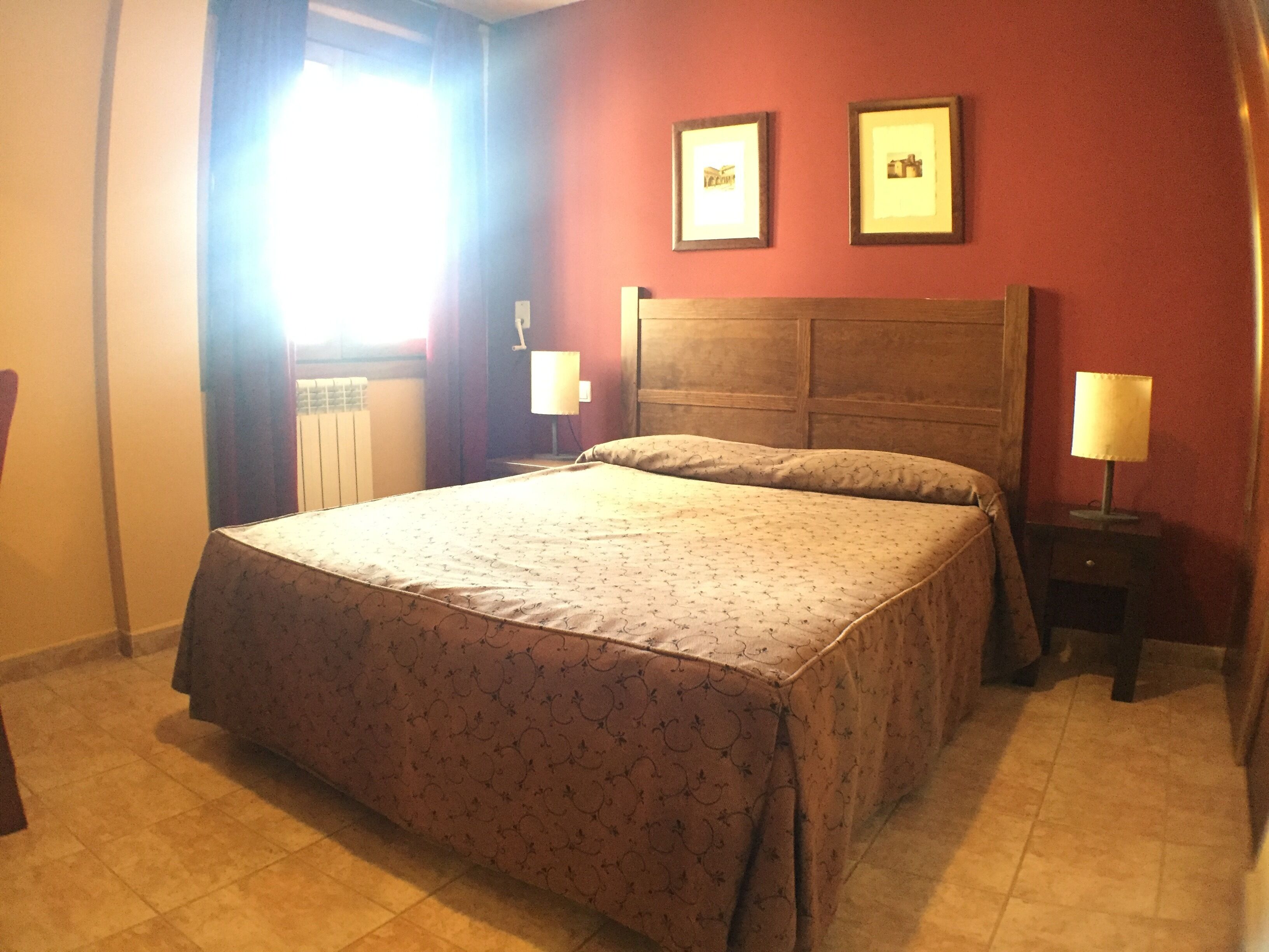 A standard hotel room with a bed, two bedside tables with lamps, a window with curtains, and two framed pictures on the wall.