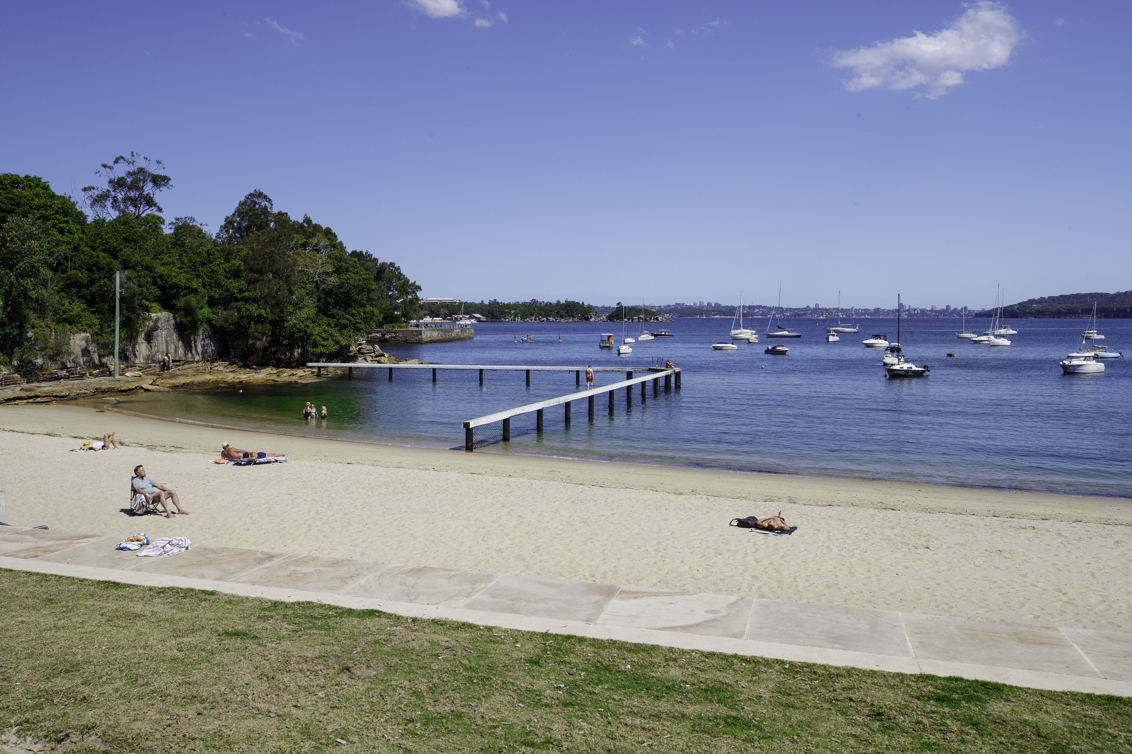 A beach with a pier extending into calm waters, surrounded by greenery and a clear sky.