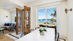 A dining area with a view of the sea and boats, a wooden door with intricate metalwork, and a white table with a vase of flowers.