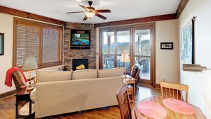 A cozy living room with a fireplace, a sofa, a dining table with chairs, and a ceiling fan. - Chalakee Lodge CI-1- Mountain View, Gated Resort, Indoor/Outdoor Pools & Hot Tub (Boone)