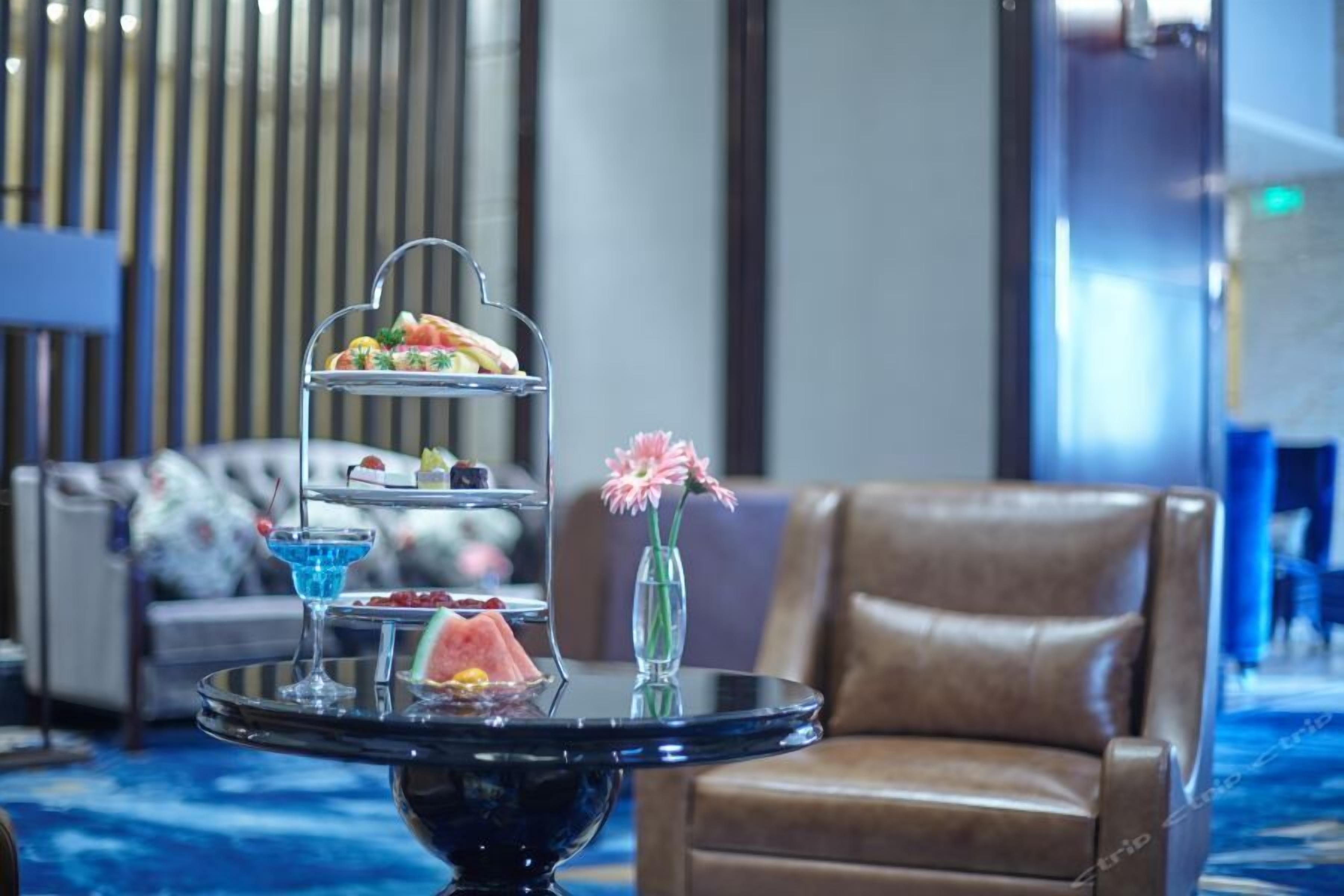 Een drielaags etagère met diverse lekkernijen en een blauwe cocktail op een ronde tafel, geplaatst in een kamer met een bruine leren stoel en een decoratieve vaas met bloemen.