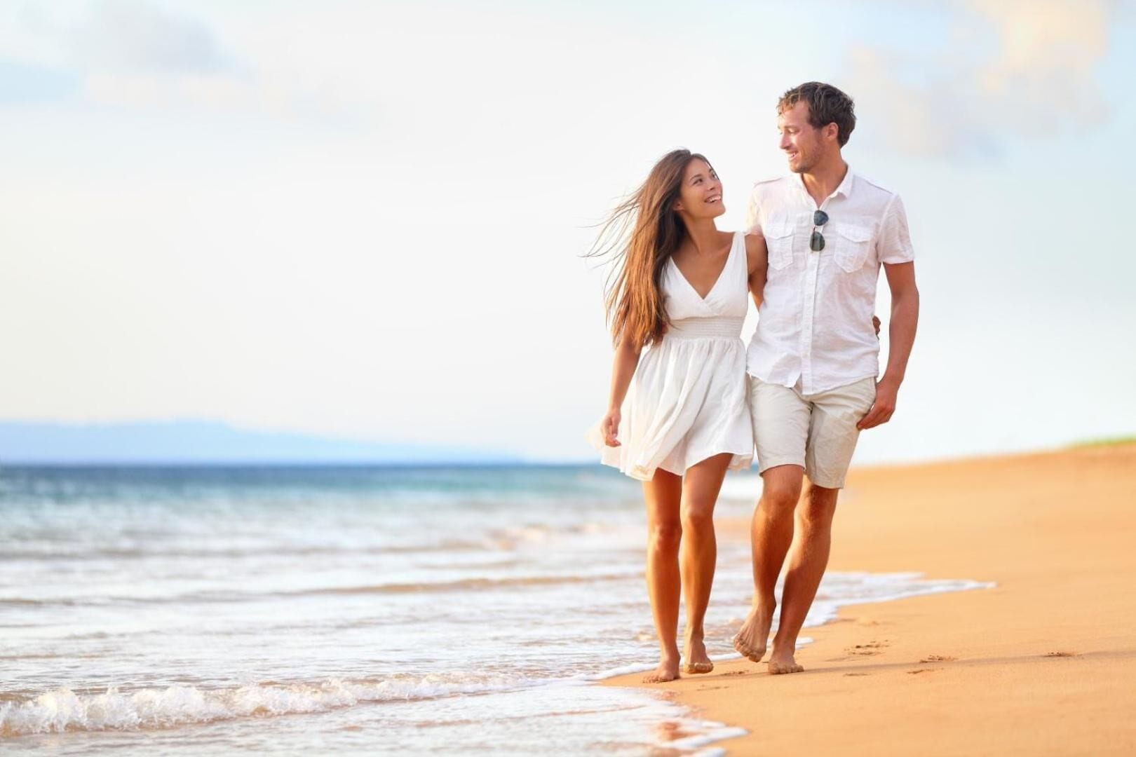 Una pareja camina sobre una playa arenosa, con el océano al fondo.