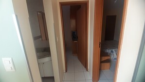 Un pasillo estrecho con una escalera a la izquierda, una puerta que da a una habitación con cama y una mesita, y suelo de baldosas.