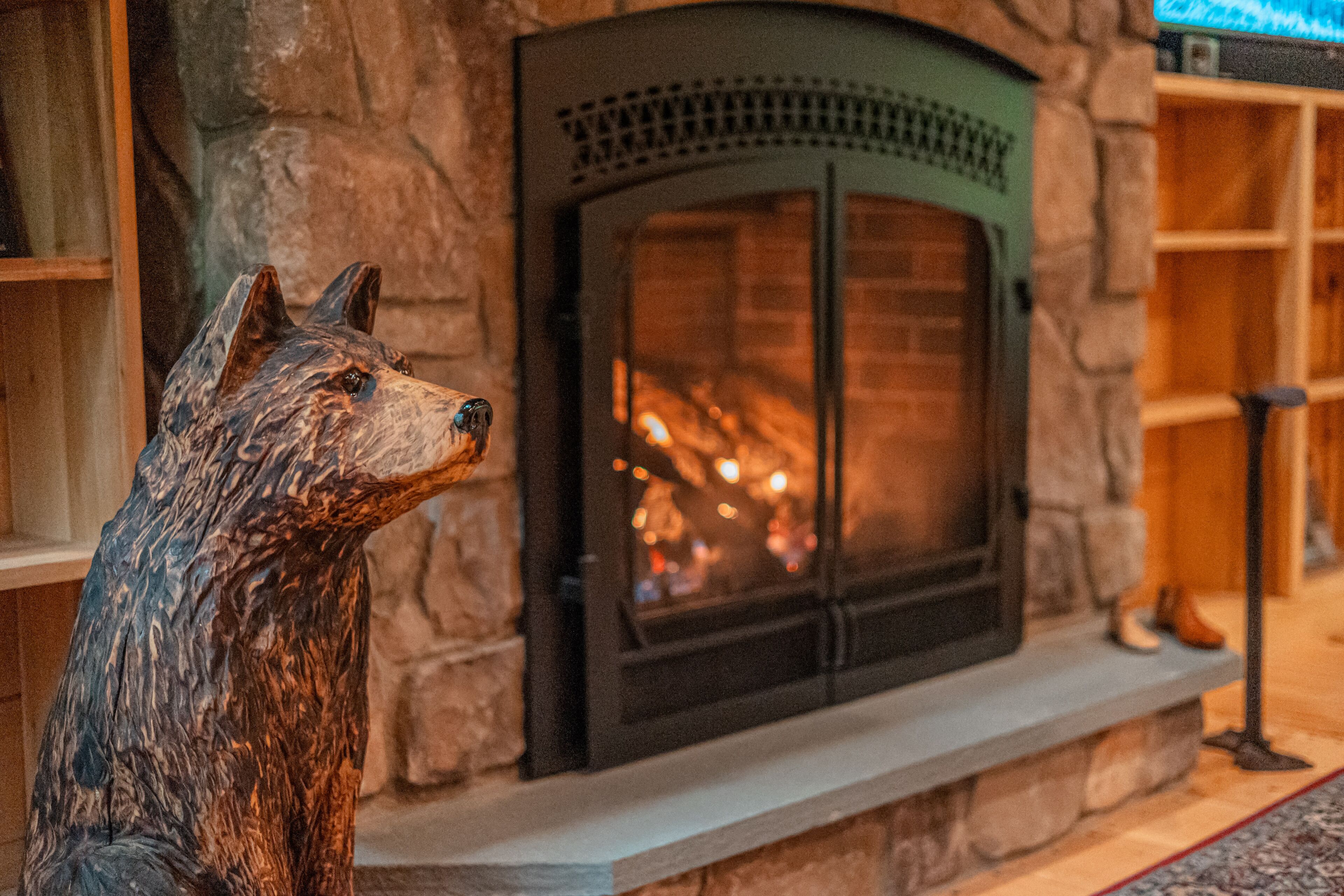 Une cheminée en pierre avec un paravent en métal, une étagère en bois sur laquelle est posée une botte, et une sculpture représentant la tête d’un loup.