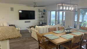 A modern living room with a large TV, a dining area with a wooden table and wicker chairs, and a view of the ocean through large windows. - Beautiful Lakefront Property with Private Dock and Beach. Renovated spring of 23 (Lake Placid)