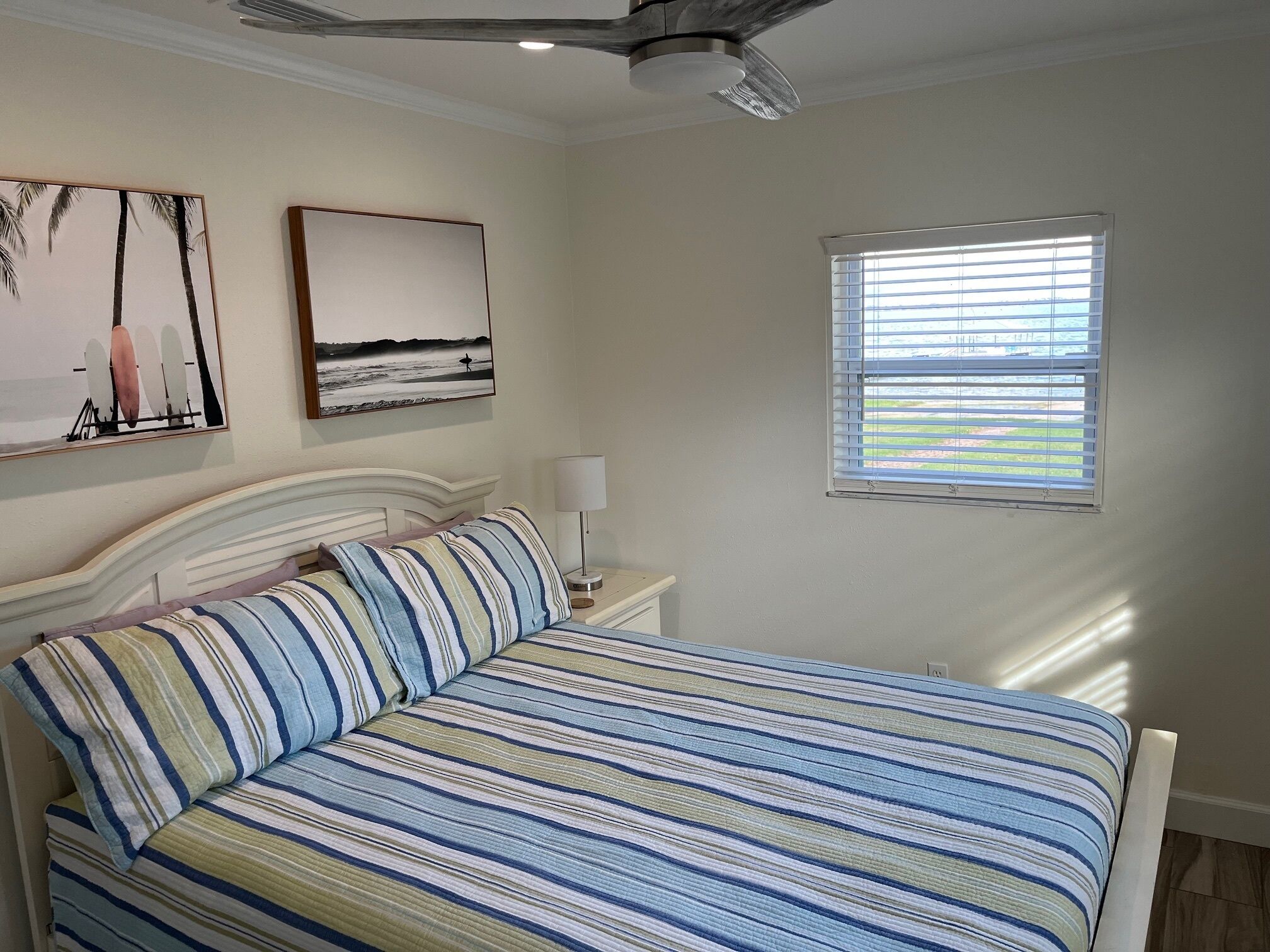 A bedroom with a bed, a nightstand, a lamp, a ceiling fan, and a window with blinds.