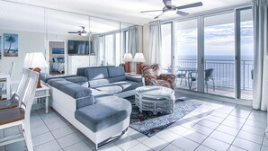 Ein modernes Wohnzimmer mit einer großen Couch, einem Esstisch und Stühlen. Ein Balkon bietet Blick auf den Ozean.