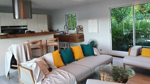 Un soggiorno moderno con un divano, poltrone e un tavolino in vetro. Si nota una zona cucina con un fornello e pensili sullo sfondo.