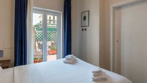 A hotel room with a bed, a door, a window with curtains, and a view of an outdoor area with plants.
