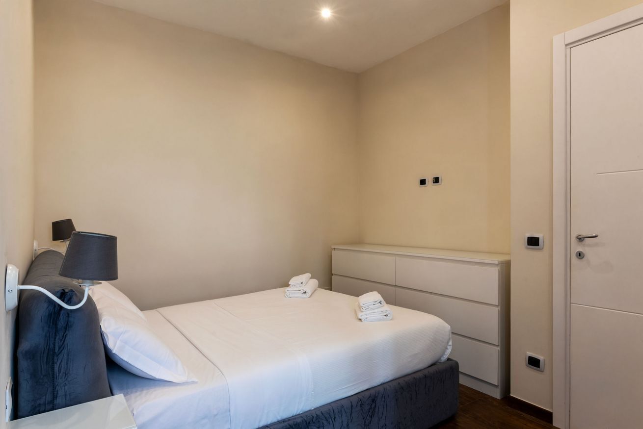 A bedroom with a bed, bedside lamp, two bedside tables, a wardrobe, and a wall-mounted light fixture.