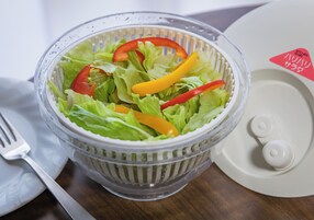 A clear plastic salad bowl with lettuce and sliced bell peppers on a wooden table.