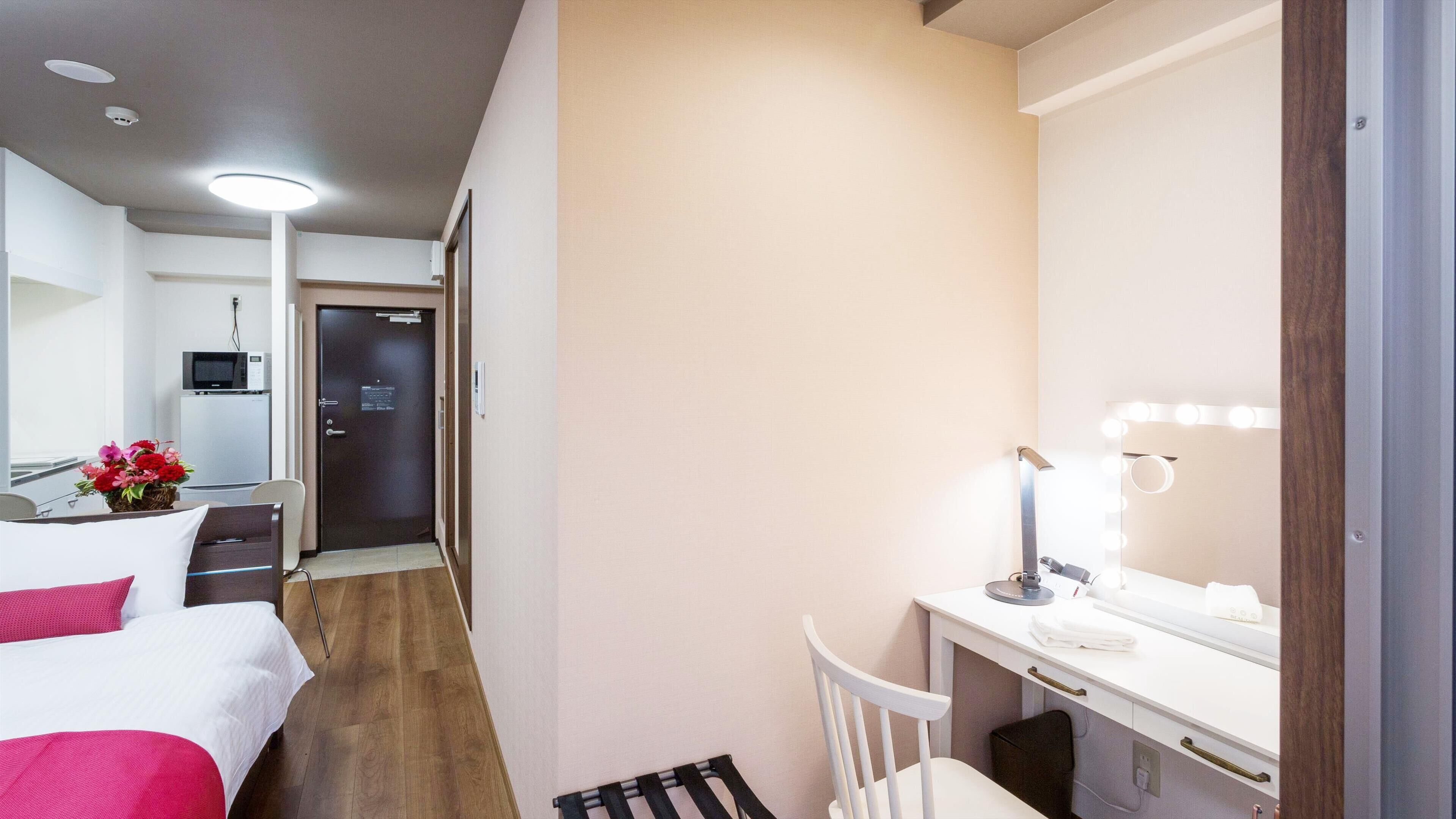 A hotel room with a bed, a desk with a mirror and lights, a chair, a small table with a vase of flowers, and a door leading to a hallway with a microwave and refrigerator.