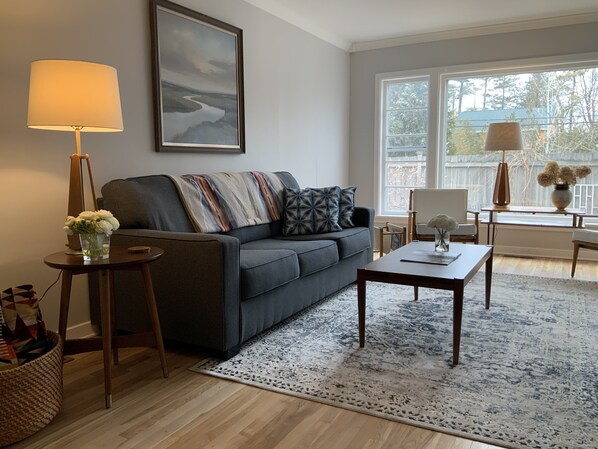 A living room with a sofa, coffee table, and lamp. There is a painting on the wall and a window with a view of trees. - Charming garden cottage downtown Ellison Bay walking distance to lake and shops (Ellison Bay)