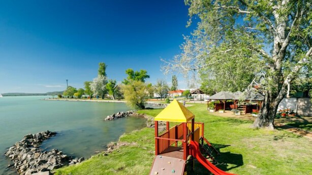 Une aire de jeux dotée d’une structure de jeu avec un toit jaune et d’un toboggan rouge, située près d’une étendue d’eau, sous un ciel dégagé.