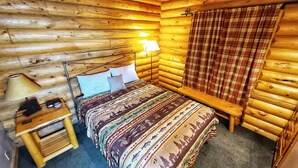 Une chambre aménagée dans un chalet en rondins, avec un lit, une lampe de chevet, une petite table et des rideaux à carreaux.