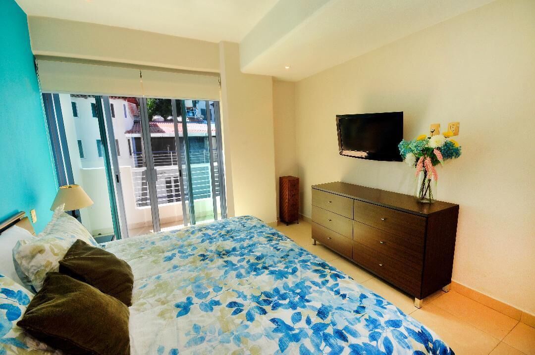 Une chambre à coucher avec un lit, une commode, un téléviseur et une fenêtre donnant sur des immeubles.