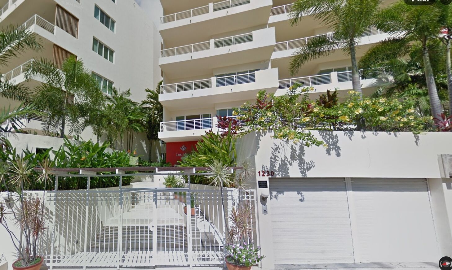 Un edificio residencial de varios pisos con balcones, entrada con portón y numerosas plantas en macetas.