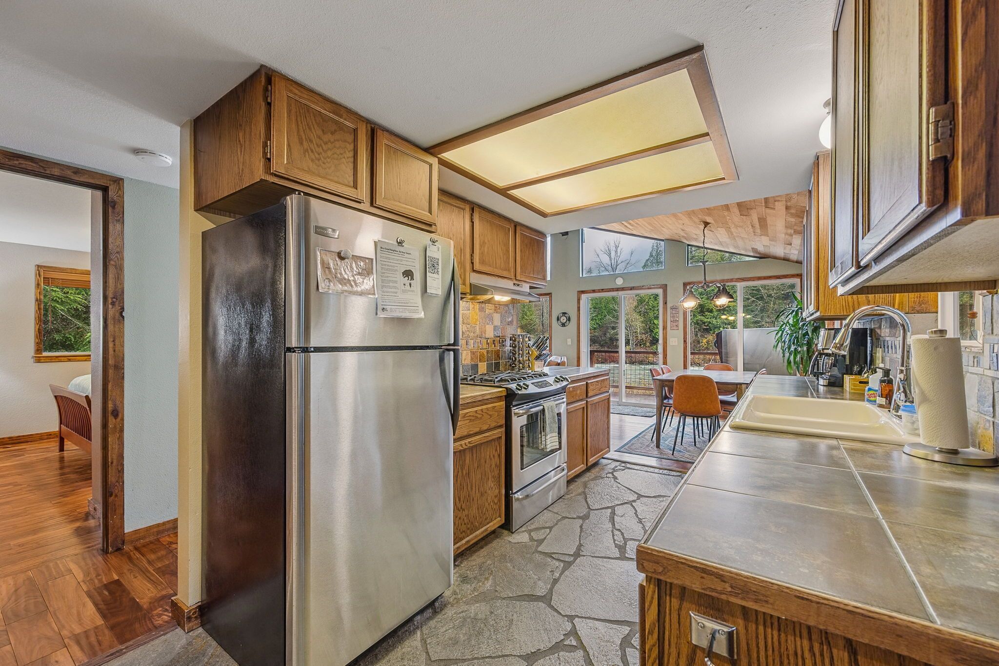 A modern kitchen with wooden cabinets, a stainless steel refrigerator, a built-in oven, and a stone tile backsplash.