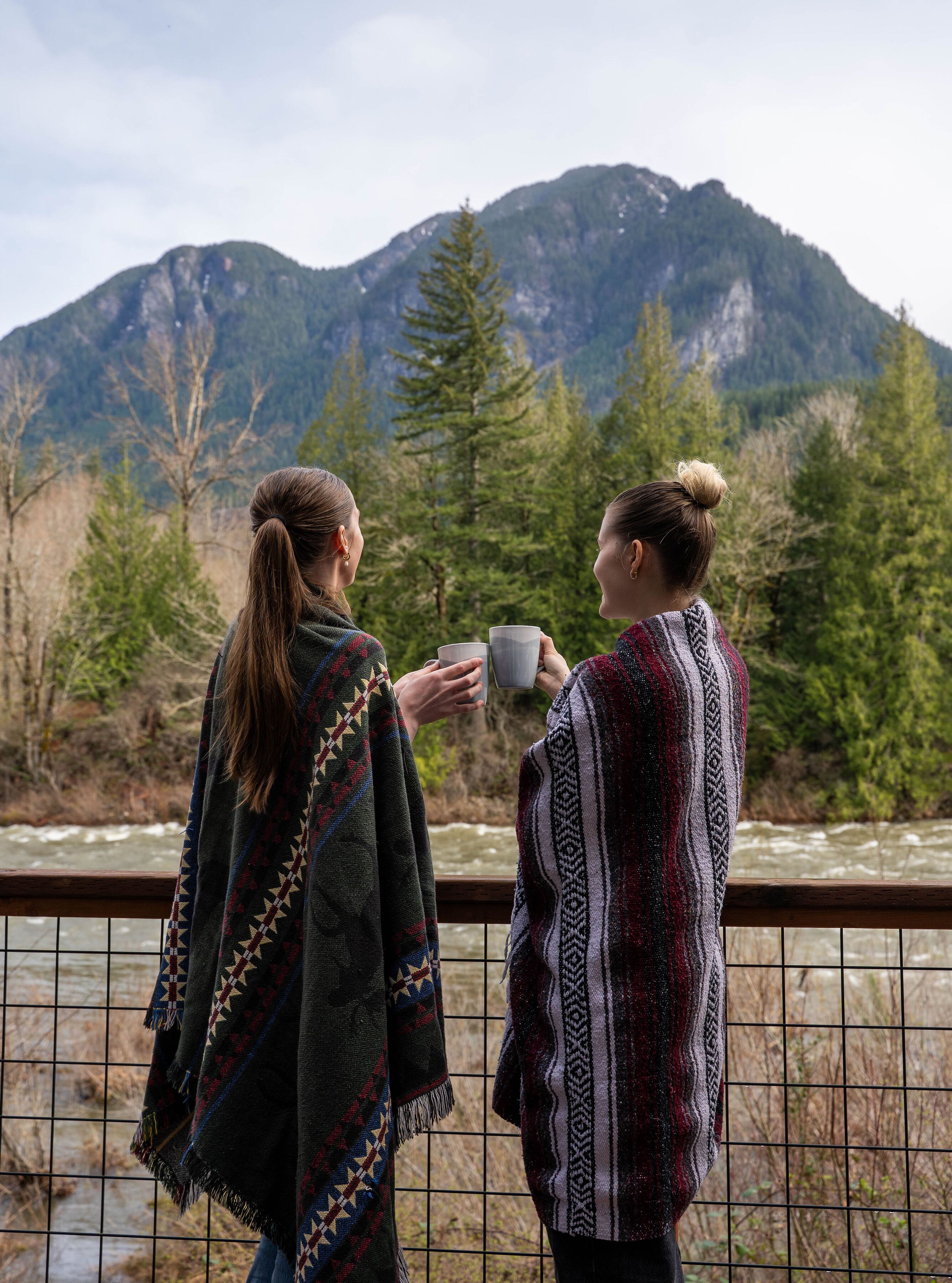 Two people standing by a railing, overlooking a river with mountains in the background.
