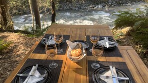 A wooden table set for four with wine glasses, plates, and a basket of bread, overlooking a flowing river.