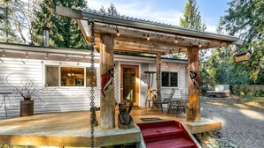 Une terrasse en bois avec un escalier rouge menant à l’entrée de la maison, le tout entouré d’arbres et bordé d’un chemin de pierres.