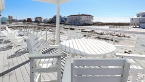 Un espace extérieur abrité, aménagé avec des chaises et des tables blanches, qui offre une vue sur une plage, avec des bâtiments visibles à l’arrière-plan.